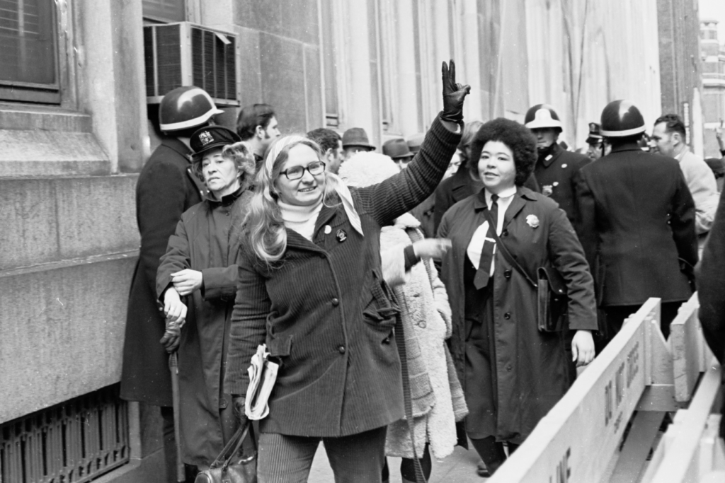 Woman giving peace sign behind police barricade followed by female officers