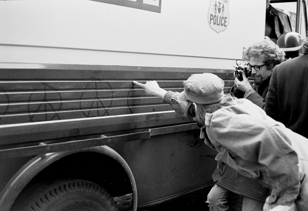 Demonstrator drawing peace sign on police van with finger while photographer shoots.